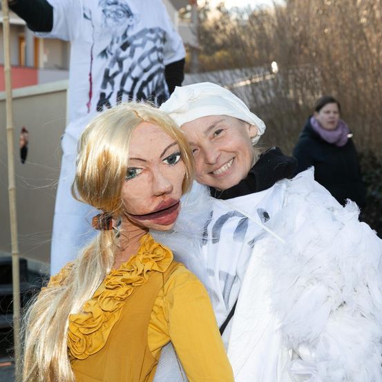 Eine blonde Frau in einem gelben Kleid und einer Maske steht neben einer Frau in einem weißen, fedrigen Kostüm. Sie lächeln. Eine Person in einer schwarzen Jacke steht hinter ihnen.