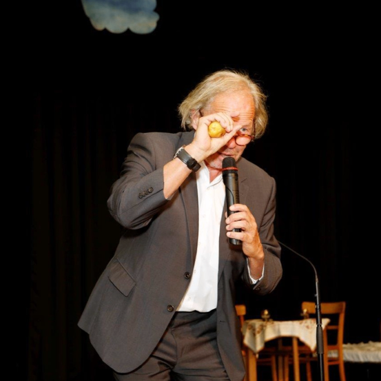 Ein älterer Mann in einem Anzug hält eine Zitrone in einer Hand und ein Mikrofon in der anderen und steht auf der Bühne.