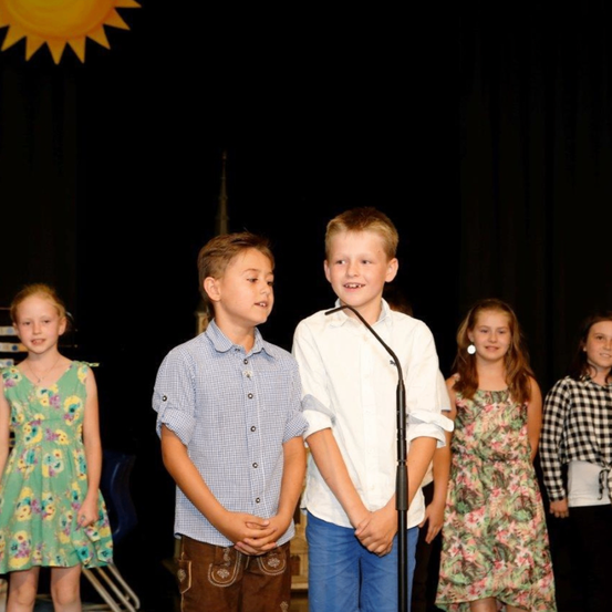 Fünf Kinder, zwei Jungen und drei Mädchen, stehen auf einer Bühne mit einem Mikrofon davor. Sie alle lächeln. Dahinter ist ein schwarzer Vorhang mit einem Sonnengrafik.