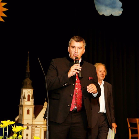 Ein Mann steht auf der Bühne, hält ein Mikrofon in der Hand, trägt eine rote Krawatte und einen schwarzen Anzug. Ein weiterer Mann steht hinter ihm. Die Bühne ist dunkel, mit einer kirchenähnlichen Struktur und einem dekorativen Hintergrund.