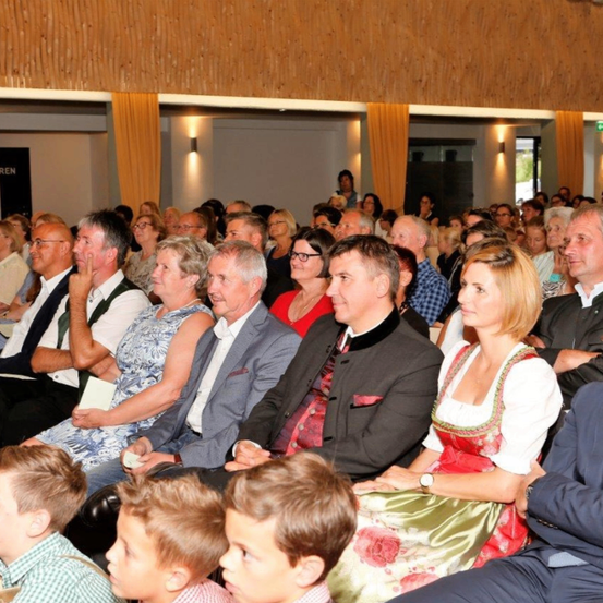 Eine große Gruppe von Menschen, darunter Erwachsene und Kinder, sitzt in einem Theater oder Konferenzsaal und hört aufmerksam einer Präsentation zu. Einige sind in formeller Kleidung, andere in lässiger Kleidung gekleidet. Der Raum hat Holzwände und Vorhänge.