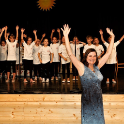 Eine Frau steht mit erhobenen Händen auf der Bühne, umgeben von Kindern in weißen Shirts und schwarzen Hosen. Die Kinder heben ihre Hände in die Luft.