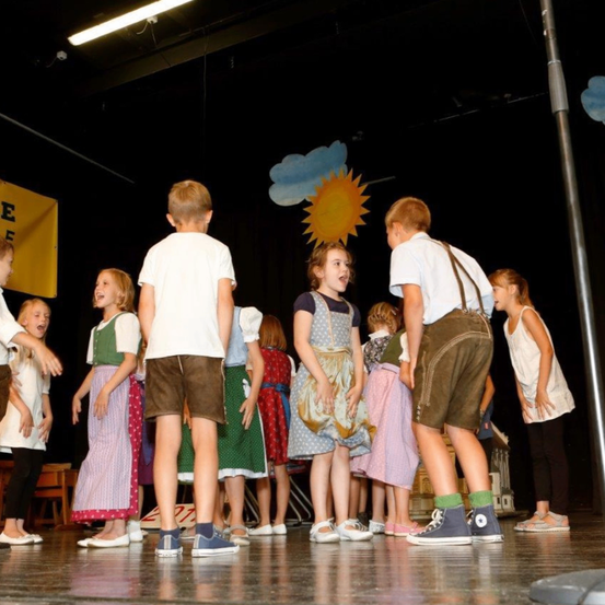 Eine Gruppe von Kindern in traditioneller Kleidung tritt auf der Bühne auf. Die Bühne hat einen Hintergrund mit Sonne und Wolken und ein gelbes Banner mit dem Wort E.