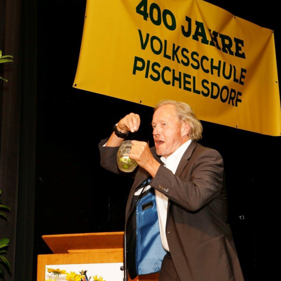 Ein Mann in einem Anzug hält ein Glas und spricht an einem Podium mit einem Banner hinter ihm, auf dem 400 Jahre Volksschule Pischelsdorf steht.