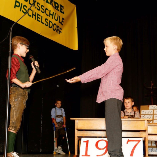 Zwei Kinder auf der Bühne, eines hält einen Stock und ein Mikrofon, das andere gestikuliert. Dahinter ein Junge mit Krücken. Ein Tisch mit Nummern. Ein Banner mit 'PISCHELSCHULE PISCHELSDORE'.