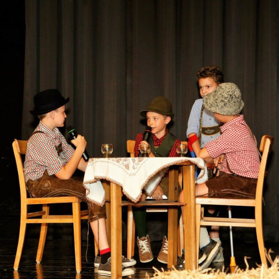 Vier Jungen sitzen um einen Tisch herum, jeder hält ein Mikrofon. Ein Junge trägt einen Hut und alle sind in traditioneller Kleidung gekleidet. Sie führen wahrscheinlich auf der Bühne auf.