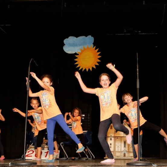 Eine Gruppe von Kindern in gelben T-Shirts und schwarzen Hosen tritt auf der Bühne auf. Zwei Mädchen springen in der Luft, während andere dahinter stehen. Ein Mikrofon steht auf der Bühne.