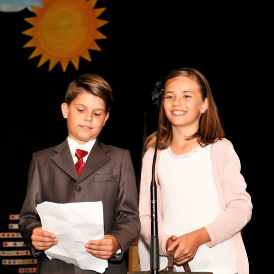 Ein Junge in einem Anzug liest von einem Stück Papier, während ein Mädchen neben ihm lächelt. Dahinter befindet sich eine Kulisse einer Sonne.