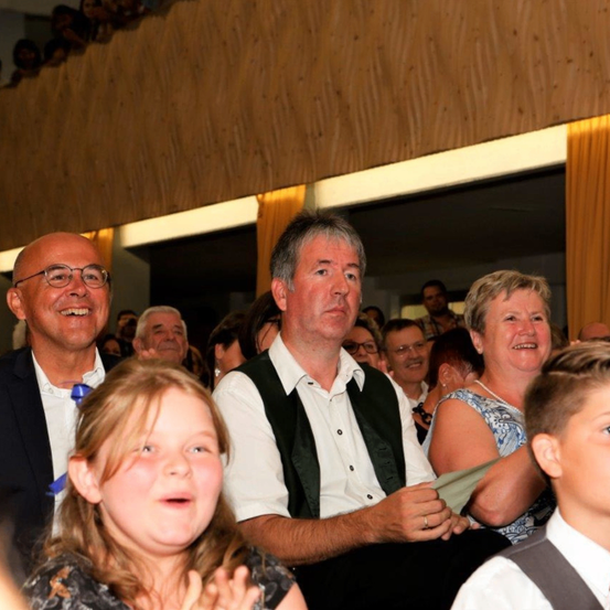 Eine Gruppe von Menschen sitzt in einem Auditorium, einige lächeln und andere schauen aufmerksam zu. Das Publikum besteht aus Männern und Frauen sowie Kindern. Die Umgebung scheint ein Indoor-Event zu sein.