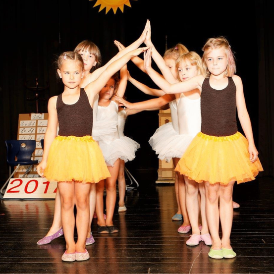 Mehrere junge Mädchen führen eine Tanzroutine auf einer Bühne auf. Die Mädchen tragen passende gelbe Tüllröcke und braune Tanktops. Sie stehen in einer Reihe mit erhobenen Händen. Dahinter steht eine Holzkiste mit Zahlen und ein Stuhl.