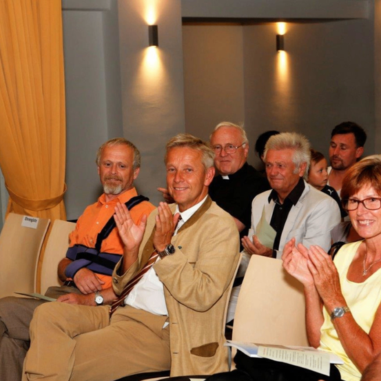 Mehrere Erwachsene sitzen in einem Raum und klatschen. Der Mann in der Mitte trägt eine helle Jacke. Ein Mann rechts trägt einen schwarzen Talar. Zwei Lampen sind an der Wand montiert.
