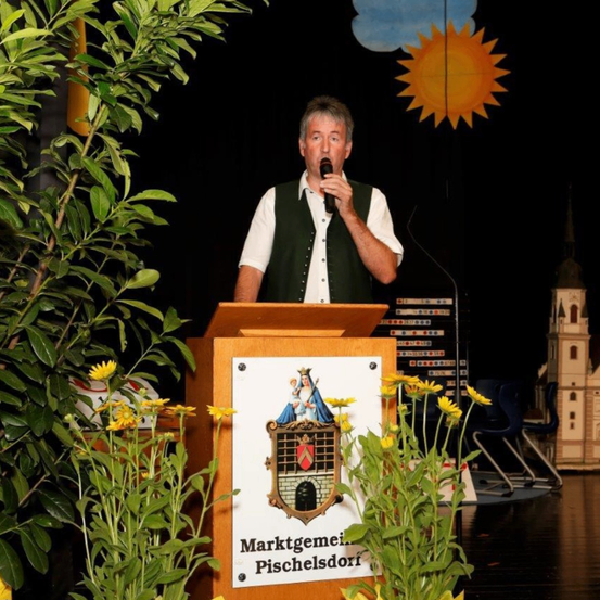 Ein Mann steht an einem Rednerpult vor einer Kulisse mit Sonne und Kirche, trägt eine Weste. Er hält ein Mikrofon und scheint eine Rede zu halten.