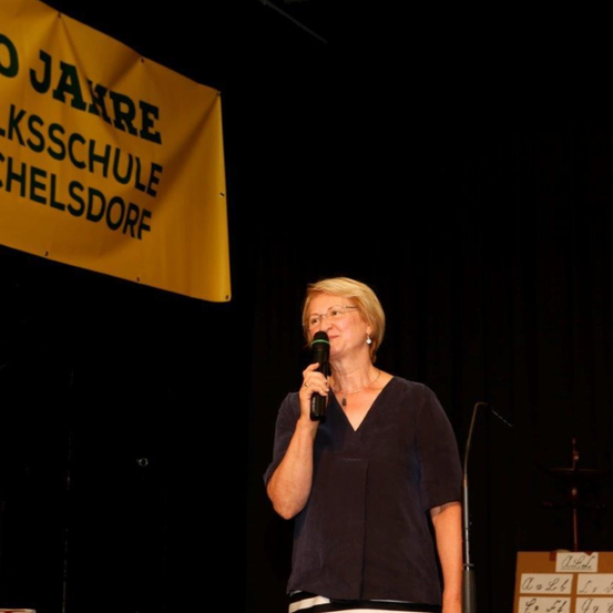 Eine Frau steht auf der Bühne, hält ein Mikrofon und spricht hinein. Sie trägt eine Brille und ein dunkles Oberteil. Ein Banner hinter ihr lautet 'O JAHRE LKSCHULE CHELSDORF'. In der Nähe befindet sich ein Mikrofonstativ und ein kleines Tafel mit Zahlen.