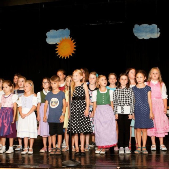 Eine Gruppe von Kindern, einige in Kleidern und andere in Freizeitkleidung, steht auf der Bühne vor einem Mikrofon. Hinter ihnen befindet sich eine Kulisse mit Sonne und Wolken.