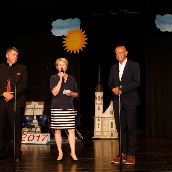 Drei Personen stehen auf einer Bühne mit Hintergrund von Sonne, Wolken und einer Kirche. Die Frau spricht in ein Mikrofon. Ein Mann mit Brille lächelt. Ein Schild auf dem Boden zeigt 2017.