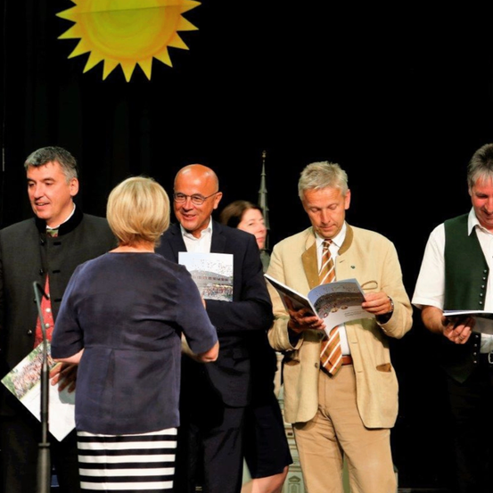 Fünf Personen stehen auf einer Bühne und halten Bücher. Im Hintergrund befindet sich eine große gelbe Sonne. Der Mann in der Mitte liest ein Buch, während die anderen ihn ansehen.
