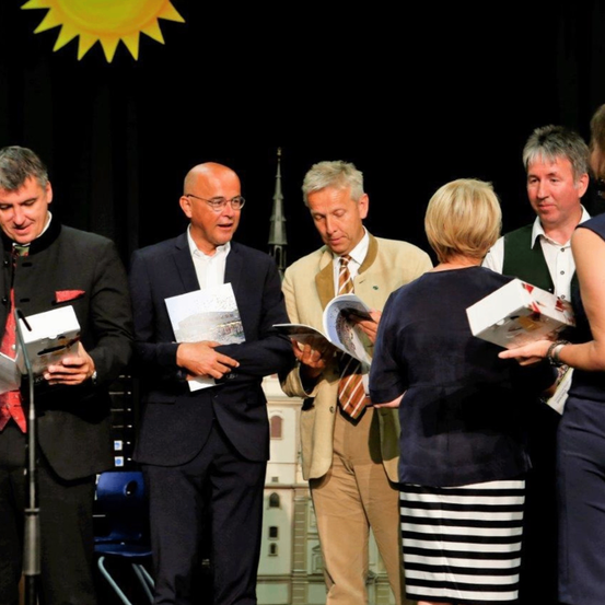 Mehrere Personen stehen auf einer Bühne und halten Bücher und Papiere in den Händen, möglicherweise bei einer Preisverleihung. Im Hintergrund ist ein Hintergrund mit einer Sonne und einem Gebäude zu sehen.