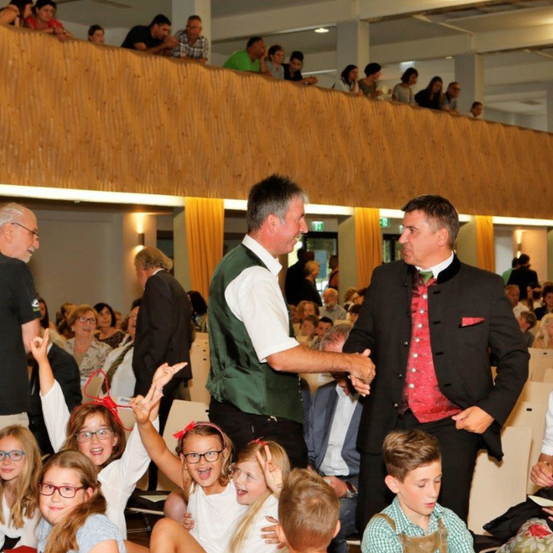 Zwei Männer in traditioneller Kleidung schütteln sich die Hände in einem überfüllten Raum. Kinder und Erwachsene sitzen und stehen, einige beobachten. Der Raum hat ein hölzerne Gesims, Vorhänge und Deckenleuchten.