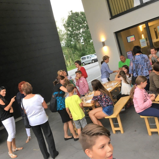 Eine Gruppe von Menschen, einschließlich Kindern, versammelt sich um Tische vor einem Gebäude. Sie scheinen eine Mahlzeit einzunehmen. Einige stehen, andere sitzen auf Bänken. Bäume und ein geparktes Auto sind im Hintergrund zu sehen.