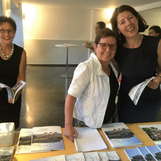 Drei Frauen stehen hinter einem Tisch mit Broschüren. Sie alle lächeln. Eine Frau trägt ein weißes Hemd und eine Brille. Eine andere Frau trägt ein schwarzes Kleid. Die dritte Frau trägt ein schwarzes Kleid und eine Halskette. Hinter ihnen ist ein verschwommener Hintergrund eines Raumes mit Tischen und Stühlen.