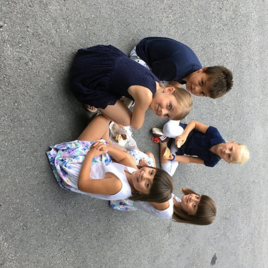Fünf Kinder sitzen im Kreis auf dem Boden und lächeln für ein Foto. Jedes Kind hält einen Snack oder ein Lebensmittel. Der Boden ist grauer Beton.