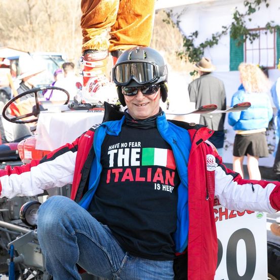 Ein lächelnder Mann in Skiausrüstung und einer Jacke mit dem Aufdruck 'Der Italiener ist hier' sitzt auf einem Fahrzeug mit einem Skistock in der Hand. Dahinter ist eine verschwommene Szene von Menschen und einem Gebäude mit Pflanzen zu sehen.