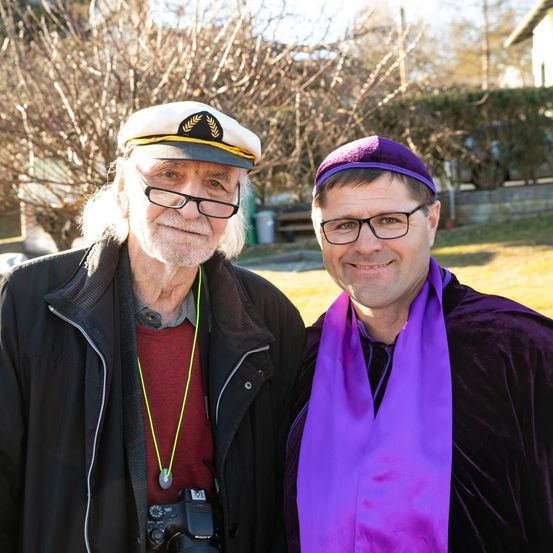 Zwei Männer lächeln und posieren für ein Foto. Der Mann auf der linken Seite trägt eine Matrosenmütze und eine Brille. Er hält eine Kamera. Der Mann auf der rechten Seite trägt einen lila Hut und einen Schal.
