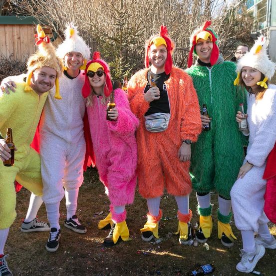 Eine Gruppe von Menschen in Hühnerkostümen posiert für ein Foto. Einige halten Flaschen. Sie stehen vor einem Gebäude mit Bäumen und Pflanzen.