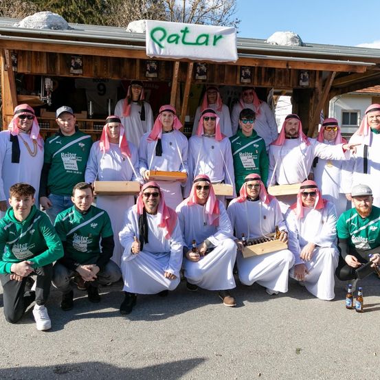 Eine Gruppe von Menschen in traditioneller arabischer Kleidung posiert für ein Foto vor einem Laden mit dem Schild 'Qatar' oben. Sie halten Kisten und Flaschen Bier.