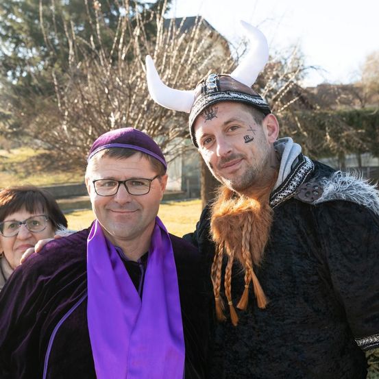 Zwei Männer in Kostümen posieren für ein Foto. Ein Mann trägt eine lila Schürze und eine Brille, der andere einen gehörnten Helm und einen Bart. Eine Frau mit einer Brille steht hinter ihnen.