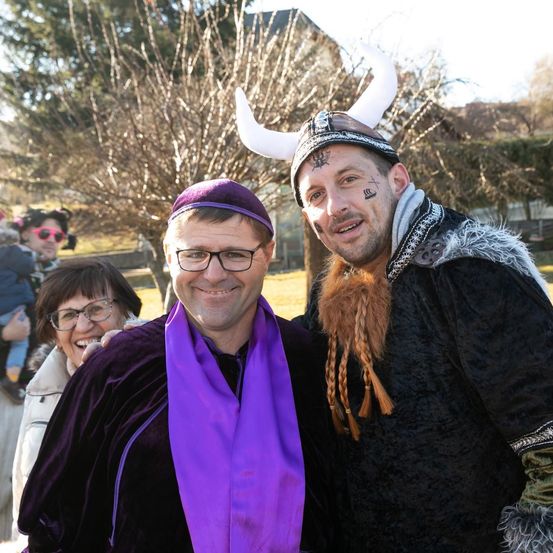 Zwei Männer in Kostümen posieren für ein Foto. Einer trägt einen Wikingerhelm und einen Bart. Der andere trägt ein lila Tuch. Eine Frau mit Brille lächelt hinter ihnen. Ein Kind ist im Hintergrund.