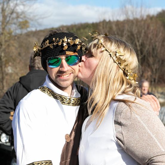 Ein Mann und eine Frau in Kostümen küssen sich, umgeben von anderen in einer natürlichen Umgebung.