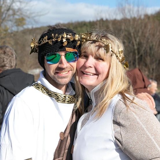 Ein Mann und eine Frau in Kostümen, lächeln für die Kamera mit Kränzen auf dem Kopf, stehen in einem Außenbereich.