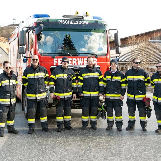 Eine Gruppe von Feuerwehrleuten in Uniform vor einem Feuerwehrauto mit dem Wort 'Feuerwehr' darauf.