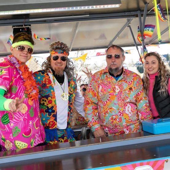 Vier Personen in leuchtenden, bunten Kostümen stehen hinter einer Bartheke und lächeln in die Kamera. Flaggen und ein Friedenssymbol sind im Hintergrund zu sehen.