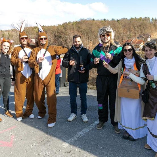 Eine Gruppe von Menschen in Kostümen, zwei in Känguruskostümen, posiert für ein Foto mit Getränken in den Händen. Dahinter ist ein bewaldeter Bereich.
