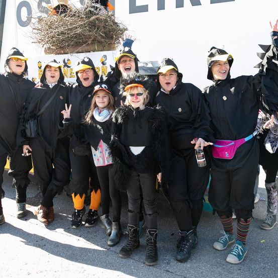 Eine Gruppe von Menschen in schwarzen Kostümen mit gelben Schnäbeln und Brillen posiert für ein Foto. Eine Person hält eine Flasche und trägt eine rosa Tasche. Sie stehen auf einer Betonfläche.