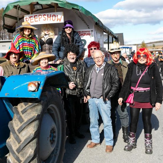 Eine Gruppe von Menschen in Kostümen, einige mit Perücken, posiert für ein Foto vor einem Traktor mit einem 'Deepshow'-Banner. Dahinter steht ein Gebäude mit einem Schild für 'Chelsdor'. Der Himmel ist bewölkt.