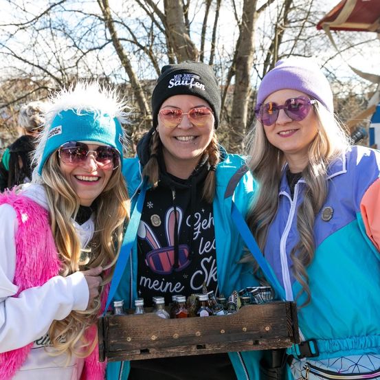 Drei Frauen, alle lächelnd, stehen zusammen und halten eine Holzkiste voller Flaschen. Sie tragen farbenfrohe Winterkleidung, einschließlich Mützen und Sonnenbrillen. Hinter ihnen deuten kahle Bäume und ein verschwommener Hintergrund auf eine Außenumgebung hin.