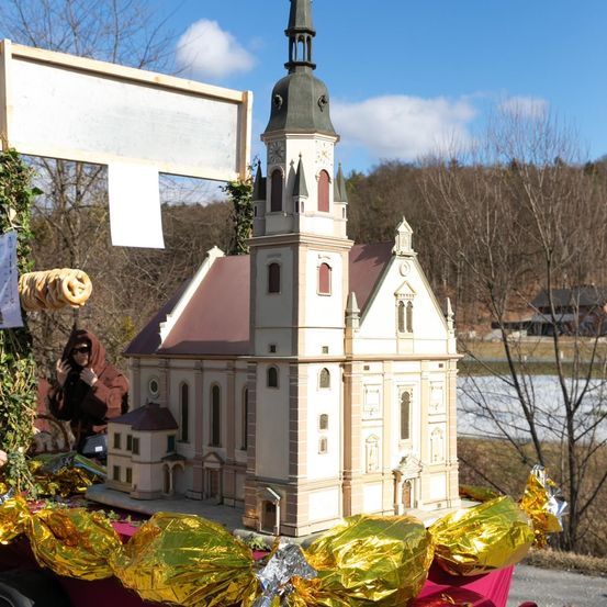 Ein kleines Modell einer Kirche mit Turm wird ausgestellt, umgeben von goldumwickelten Süßigkeiten und Grünpflanzen. Eine Person steht hinter dem Modell, und ein großes weißes Schild ist darüber. Bäume und ein Gewässer sind im Hintergrund.