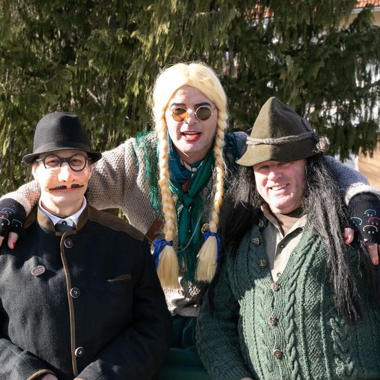 Drei Personen in Kostümen, eine mit blonden Haaren und Brille, eine weitere mit Hut und langen Haaren und die dritte mit schwarzem Hut, posieren für ein Foto.