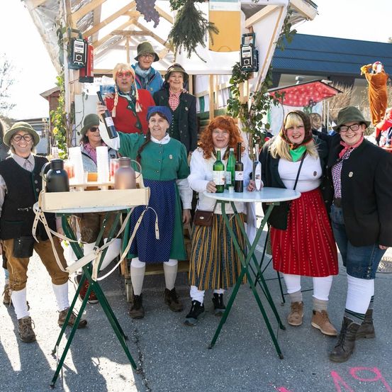 Eine Gruppe von Menschen in traditioneller Kleidung posiert für ein Foto vor einem festlichen Stand. Der Stand hat Tische mit Flaschen und Bechern. Dahinter steht ein dekorierter Stand mit Dach und einer Statue.
