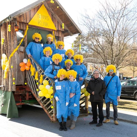 Eine Gruppe von Menschen in blauen Kostümen mit gelben Perücken posiert vor einem dekorierten Holzhaus. Das Haus trägt ein Schild mit der Aufschrift Tennis Klinik und ist von Ballons umgeben. Zwei Männer in dunklen Mänteln stehen neben ihnen.