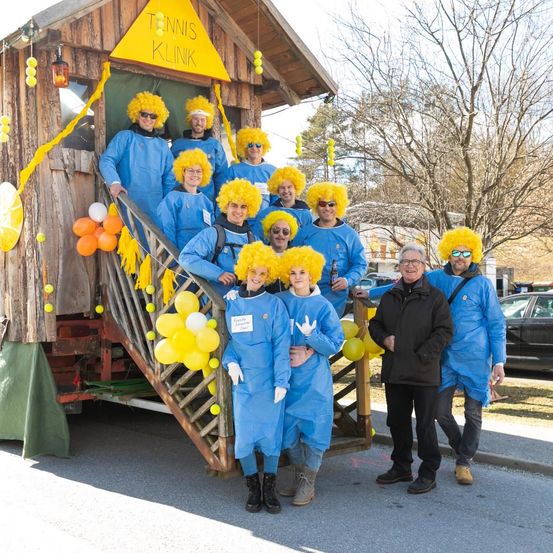 Eine Gruppe von Menschen in blauen Kitteln mit gelben Perücken posiert vor einem dekorierten Schuppen mit den Worten Tennis Klinik darauf.
