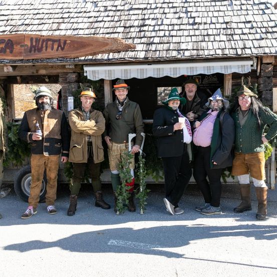 Eine Gruppe von Personen in traditioneller Kleidung, einschließlich Hüten und Stiefeln, steht vor einer Holzhütte mit einem Schild, das 'SCA HUT' steht. Sie posieren für ein Foto.