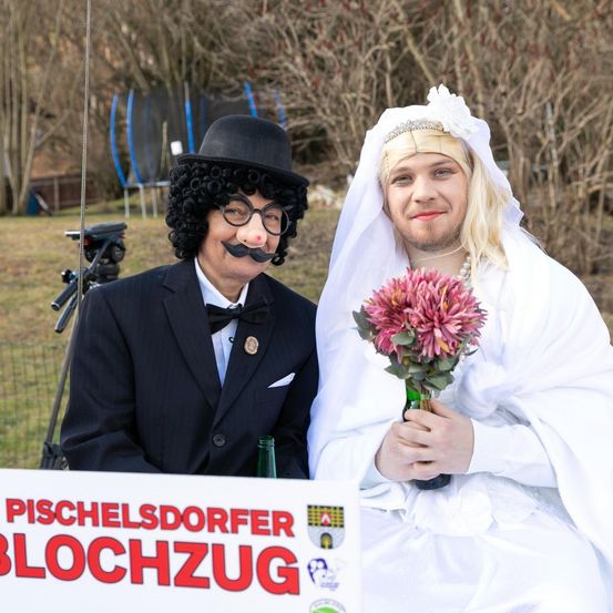 Ein Mann in einem schwarzen Anzug mit einer Schleife und einer Frau in einem Hochzeitskleid, die einen rosa Blumenstrauß hält, stehen vor einem Schild, das 'Pischelsdorfer Blochzug' besagt. Dahinter befindet sich ein Trampolin.