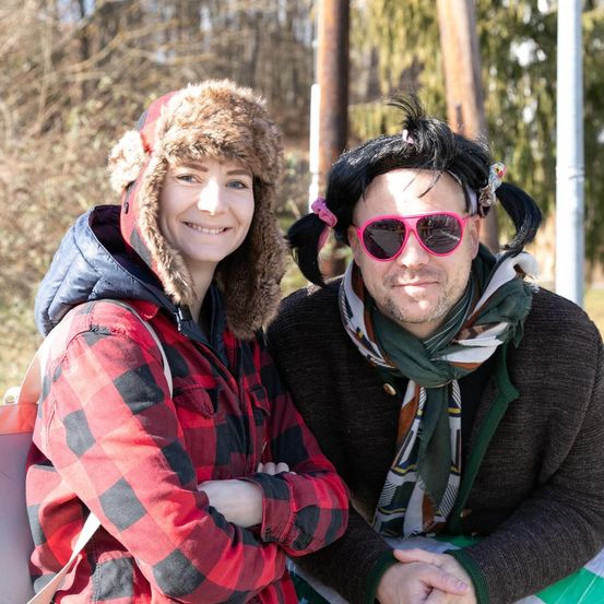 Eine lächelnde Frau in einer rot-schwarz karierten Jacke und einem Pelzhut posiert mit einem Mann in pinken Sonnenbrillen und einem Schal draußen.