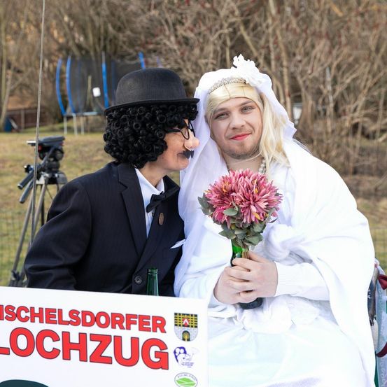 Zwei Personen in formeller Kleidung posieren für ein Foto. Einer trägt einen schwarzen Hut und eine Perücke, der andere ein weißes Schleier. Sie halten einen Strauß rosa Blumen, im Vordergrund steht ein Schild mit der Aufschrift 'SCHLESDORFER LOCHZUG'. Im Hintergrund sind ein Kamerastativ und Bäume zu sehen.