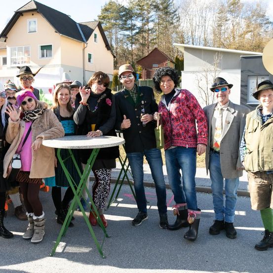 Eine Gruppe von Menschen in bunten Outfits und Hüten versammelt sich um einen Tisch vor einem Haus. Einige tragen Brillen und Stiefel. Dahinter stehen Häuser und Bäume.
