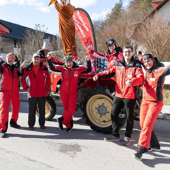 Eine Gruppe von Personen in roten und schwarzen Kleidungsstücken posiert für ein Foto vor einem roten Traktor, auf dem eine Person in einem orangefarbenen Kostüm steht.